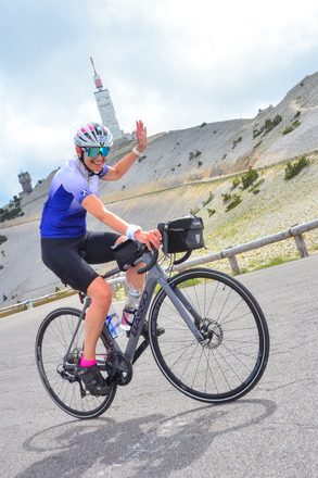 Juliet grinning and waving to the camera while cycling near the summit of Mont Ventoux, the observatory visible behind her