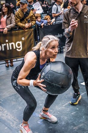 Juliet squatting with a heavy medicine ball at a HYROX fitness race, crowd watching from behind the barriers