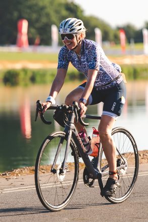 Juliet smiling as she cycles along a lakeside road in golden evening light, wearing race number 599