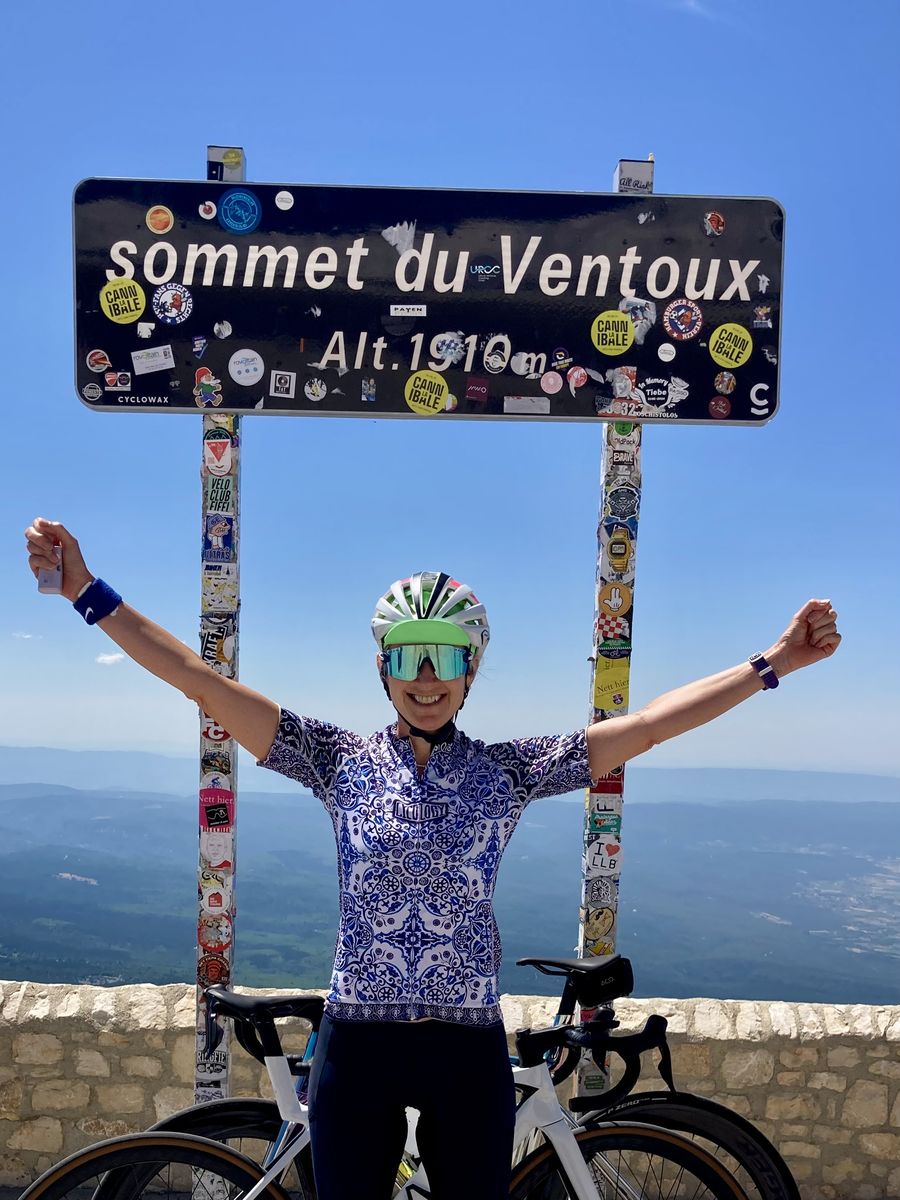 Juliet with arms raised under the summit sign at the top of Mont Ventoux, her bike beside her