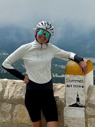 Juliet posing beside the Sommet Alt 1910m marker at the top of Mont Ventoux, storm clouds rolling in over the mountains behind her